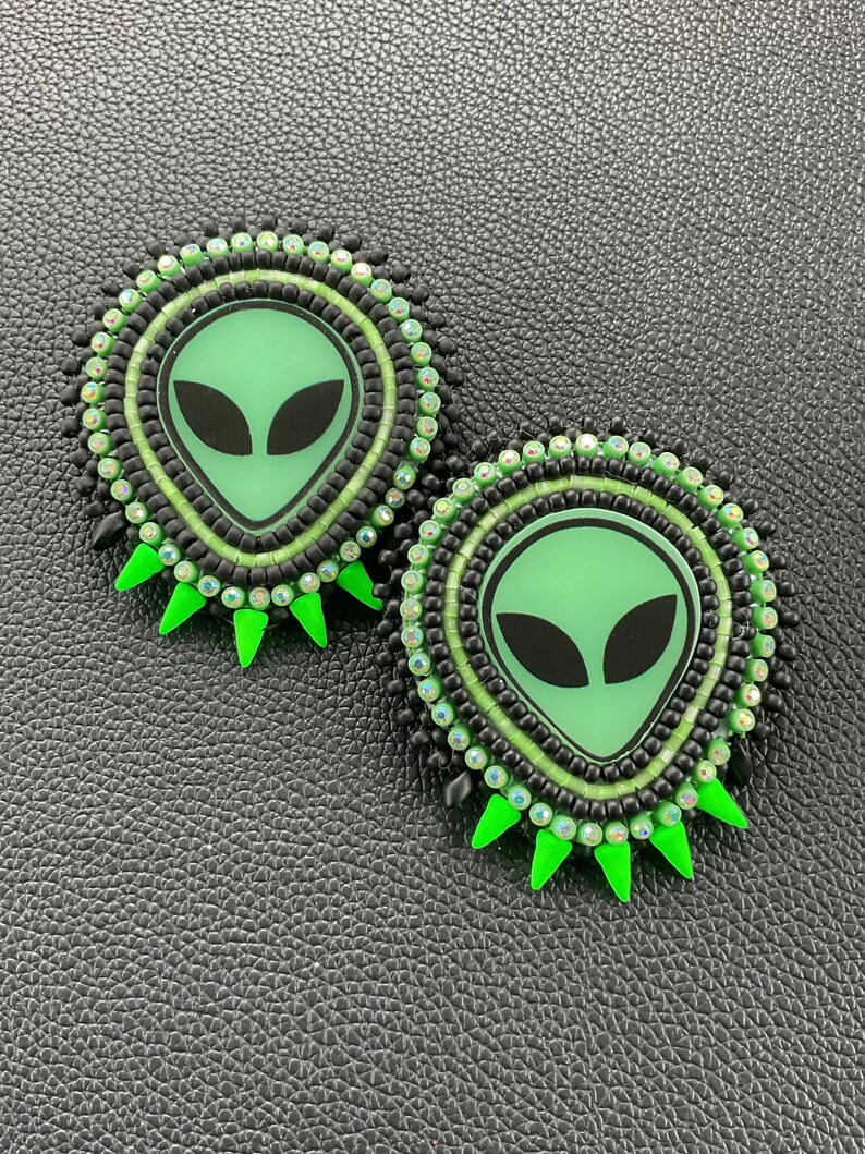 May include: Two round, beaded alien-themed earrings. Each earring features a green alien face with black eyes, surrounded by black and green beads. The earrings have green, triangular accents around the edges.