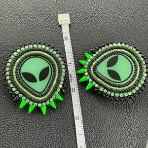 May include: Two handmade alien-themed earrings. Each earring features a green alien face with black eyes, surrounded by black and green beads. Bright green spikes extend from the outer edge. The earrings are approximately 2 inches in diameter.