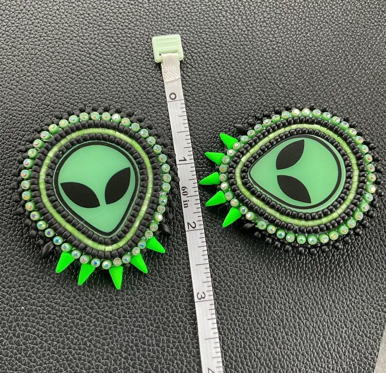 May include: Two handmade alien-themed earrings. Each earring features a green alien face with black eyes, surrounded by black and green beads. Bright green spikes extend from the outer edge. The earrings are approximately 2 inches in diameter.