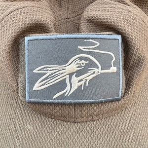 May include: A close-up of a tan baseball cap with a patch. The patch is rectangular with a light blue border and a gray background. The patch features a white embroidered design of a rabbit smoking a pipe.