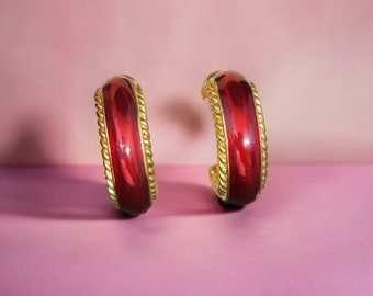 May include: A pair of gold-toned hoop earrings with a red enamel center. The earrings have a twisted rope design around the edges. The background is a gradient of pink.
