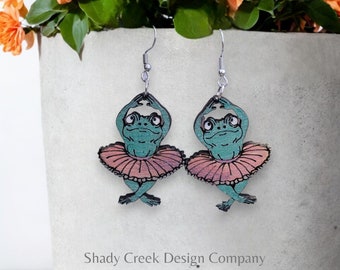 May include: A pair of dangle earrings featuring a green frog wearing a pink tutu. The frog is in a ballerina pose with its arms raised above its head. The earrings are made of wood and have silver hooks.