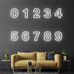 Pu&ograve; includere: Numeri bianchi al neon da 0 a 9 su una parete grigio scuro. I numeri sono contornati di bianco e hanno un bagliore rosa. Un divano giallo con un cuscino blu &egrave; in primo piano.