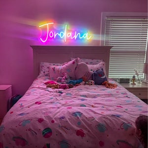 Puede incluir: Un dormitorio rosa con una cama blanca y un letrero de ne&oacute;n arco&iacute;ris que dice "Jordana" sobre la cabecera. La cama tiene una funda n&oacute;rdica estampada en rosa y blanco y est&aacute; decorada con animales de peluche.