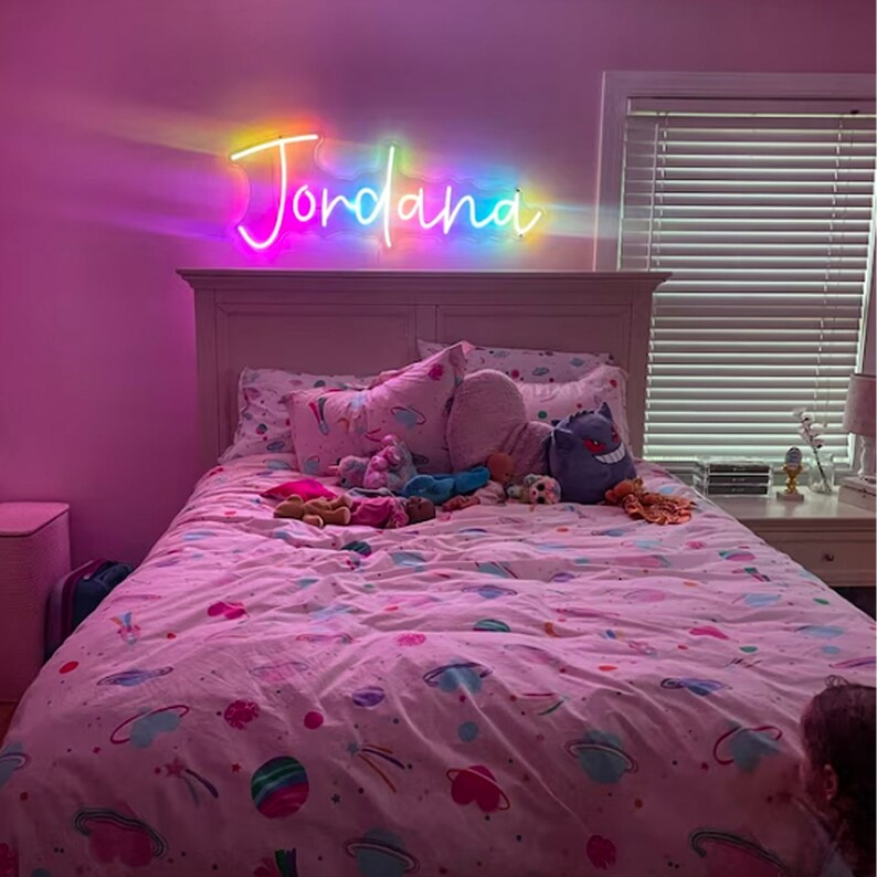 Puede incluir: Un dormitorio rosa con una cama blanca y un letrero de ne&oacute;n arco&iacute;ris que dice "Jordana" sobre la cabecera. La cama tiene una funda n&oacute;rdica estampada en rosa y blanco y est&aacute; decorada con animales de peluche.