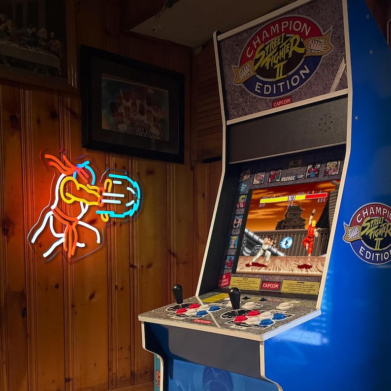 May include: A blue Street Fighter II Champion Edition arcade cabinet with a screen displaying a fighting game match. Next to it, a neon sign depicts a character performing a glowing blue energy attack, featuring orange and white light outlines.