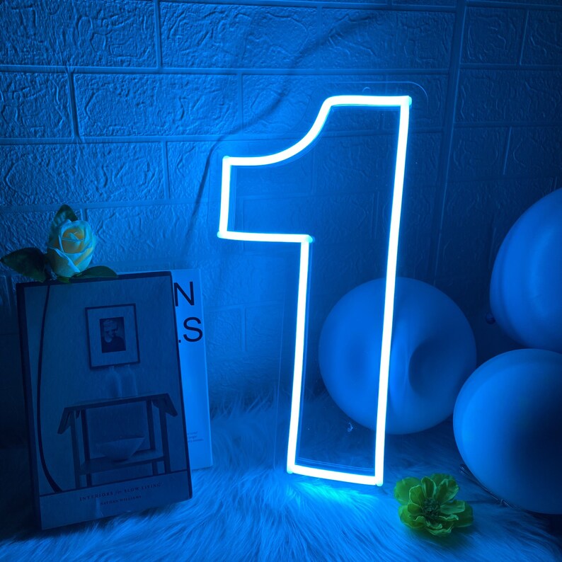 Pu&ograve; includere: Una luce al neon blu a forma di numero uno. La luce &egrave; accesa e brilla. Sullo sfondo ci sono palloncini blu.