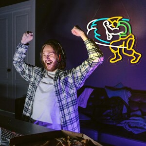 May include: A wall-mounted neon sign featuring a stylized figure in a dynamic pose with yellow, green, white, and blue light tubes. The figure is depicted with raised arms and bent legs, creating a vibrant, glowing graphic display on a dark wall.