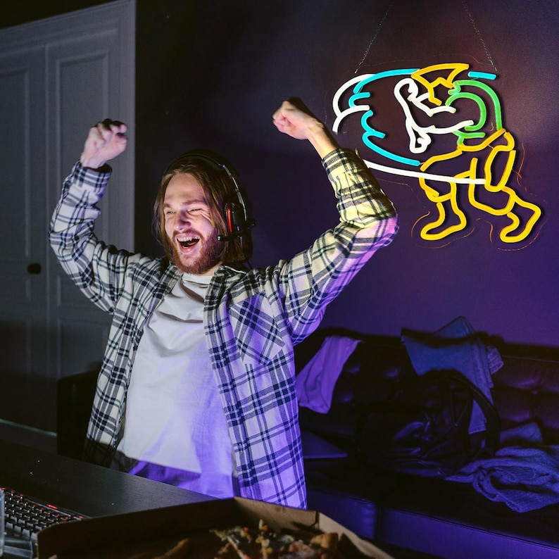 May include: A wall-mounted neon sign featuring a stylized figure in a dynamic pose with yellow, green, white, and blue light tubes. The figure is depicted with raised arms and bent legs, creating a vibrant, glowing graphic display on a dark wall.