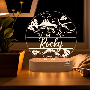 Puede incluir: Una luz nocturna de acr&iacute;lico blanco con tem&aacute;tica de dinosaurios. La luz presenta un c&iacute;rculo con un volc&aacute;n, tres dinosaurios y el nombre "Rocky" escrito debajo. La luz est&aacute; sobre una base de madera.