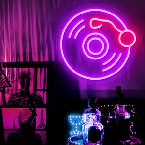 Könnte beinhalten: Ein Neonzeichen in Form einer Schallplatte leuchtet hellviolett. Das Schild ist an einer Wand angebracht, im Hintergrund befinden sich ein Fenster mit Kunstwerken und Flaschen mit Lichtern.
