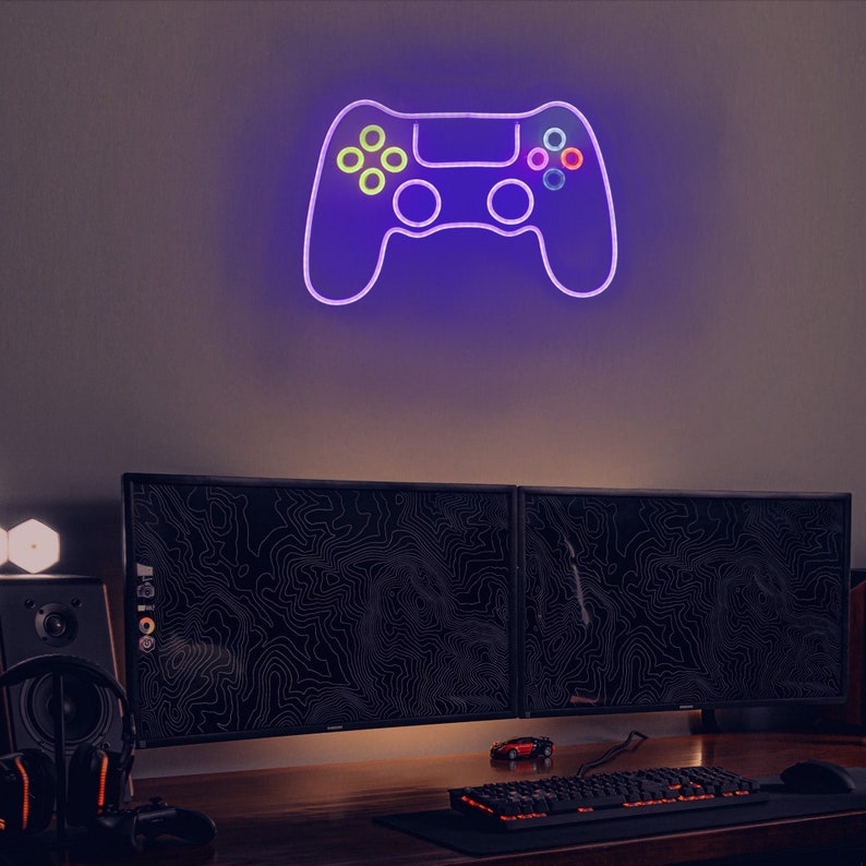 May include: A purple neon sign shaped like a video game controller with colorful buttons. The sign is mounted on a wall above two computer monitors.