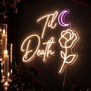 Peut inclure: Une enseigne néon affichant le texte 'Til Death' en lumière blanche chaude, accompagnée d'un croissant de lune violet et du contour d'une rose blanche. L'enseigne est fixée sur une surface sombre, avec un câble électrique sortant de la rose.