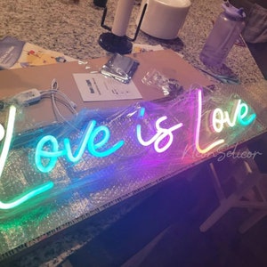 Puede incluir: Un letrero de ne&oacute;n con las palabras "Love is Love" en colores arco&iacute;ris. El letrero est&aacute; envuelto en pl&aacute;stico y est&aacute; sobre una mesa.