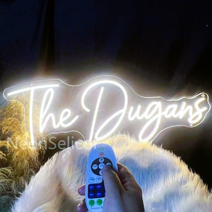 May include: A white neon sign in the shape of a guitar with the text "The Dugans" in a cursive font. A hand is holding a remote control with multiple buttons, likely to control the sign. The sign is illuminated against a dark background.