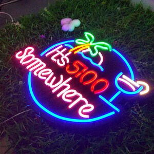 May include: Neon sign with a blue, pink and red outline. The sign says "It's 5 O'Clock Somewhere". There is a palm tree and a cocktail glass in the design.