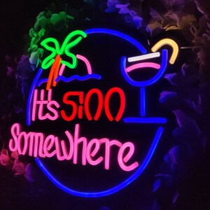 May include: Neon sign with blue, pink and red lettering that reads "It's 5:00 Somewhere". The sign also features a neon image of a palm tree and a cocktail glass.
