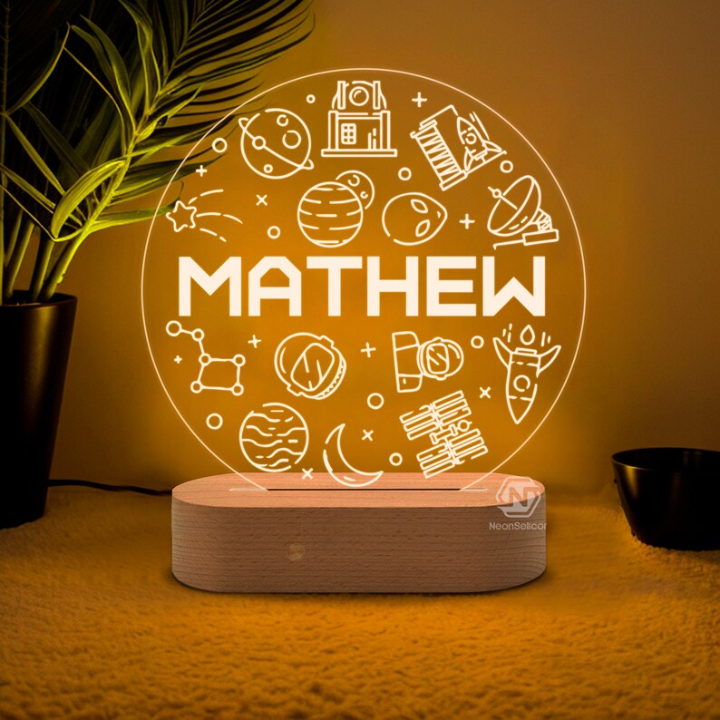 Puede incluir: Una luz nocturna LED blanca con una base de madera. La luz presenta un dise&ntilde;o circular con tem&aacute;tica espacial, que incluye planetas, estrellas, cohetes y un astronauta. El nombre "MATHEW" est&aacute; escrito en el centro del dise&ntilde;o.