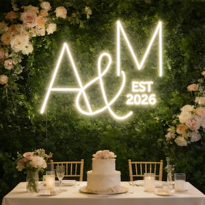 Puede incluir: Escena de recepción de boda con una tarta de dos pisos, arreglos florales y un letrero de neón que muestra las iniciales "A&M" y "EST 2026" contra una pared verde. La mesa está puesta con copas y velas.