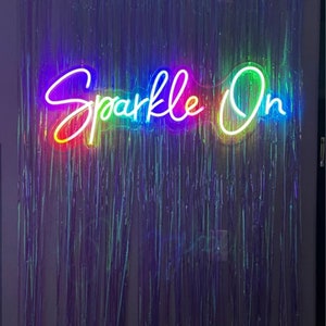 Puede incluir: Un letrero de ne&oacute;n con las palabras "Sparkle On" en colores arco&iacute;ris. El letrero est&aacute; montado en una pared con una cortina plateada iridiscente detr&aacute;s.