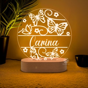 Puede incluir: Una luz nocturna de acr&iacute;lico blanco con un dise&ntilde;o circular que presenta mariposas, flores y el nombre "Carina". La luz est&aacute; sobre una base de madera.