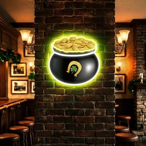 Könnte beinhalten: Ein Neonschild mit einem schwarzen Topf voller Goldmünzen, einem Hufeisen und einem Kleeblatt. Das Schild ist grün umrandet und an einer Backsteinmauer angebracht, passend zum St. Patrick's Day.