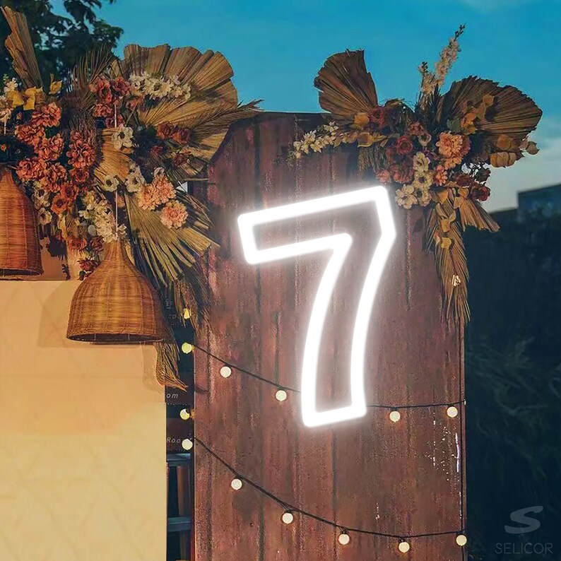 Pu&ograve; includere: Un numero sette al neon bianco su uno sfondo di legno con un'arrangiamento floreale e luci a catena. Il numero &egrave; illuminato e lo sfondo &egrave; marrone.