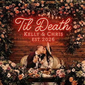 Op de afbeelding: Een rood neonbord met de tekst Til Death, KELLY & CHRIS en EST. 2026, gemonteerd op een donkere houten muur. Het bord is omringd door een weelderig arrangement van oranje, roze en witte bloemen en groen, dat een zittend paar omlijst.