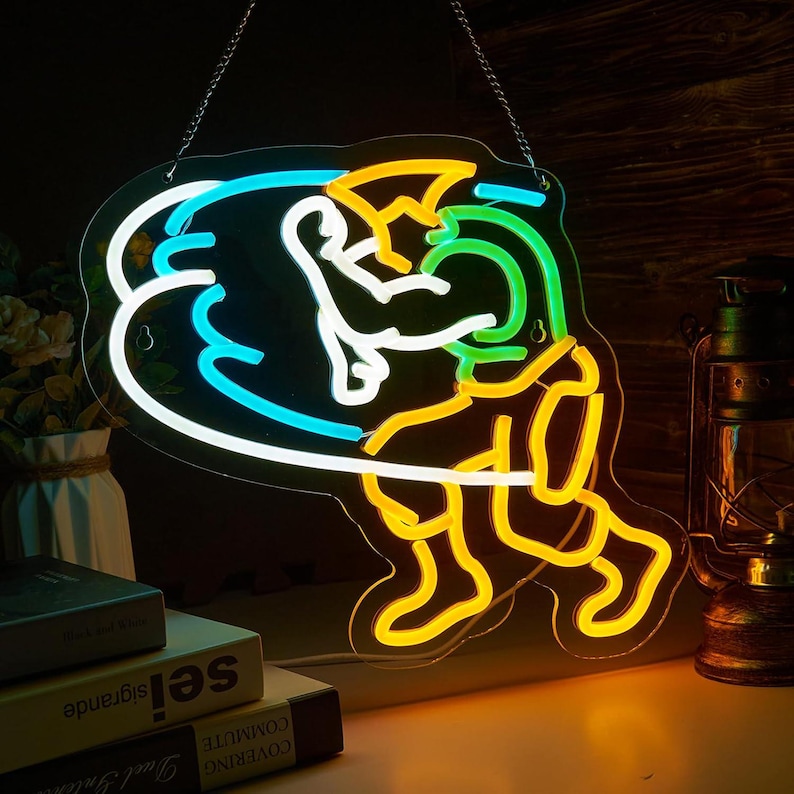 May include: A neon sign featuring a stylized figure in a dynamic pose, outlined in bright yellow, green, and blue light. The sign is suspended by a metal chain and displays a glowing, abstract character design against a dark background.