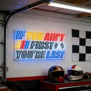 May include: A neon sign with the text "IF YOU AIN'T FIRST YOU'RE LAST" in blue, yellow, and red. Two racing helmets, one black and gold, the other white and red, sit below the sign. A checkered flag hangs in the background.
