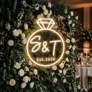 Pode incluir: Uma placa de néon em forma de anel de diamante com as iniciais "S & T" e o ano "Est. 2026". A placa é rodeada por rosas brancas e vegetação, perfeita para um casamento ou noivado.