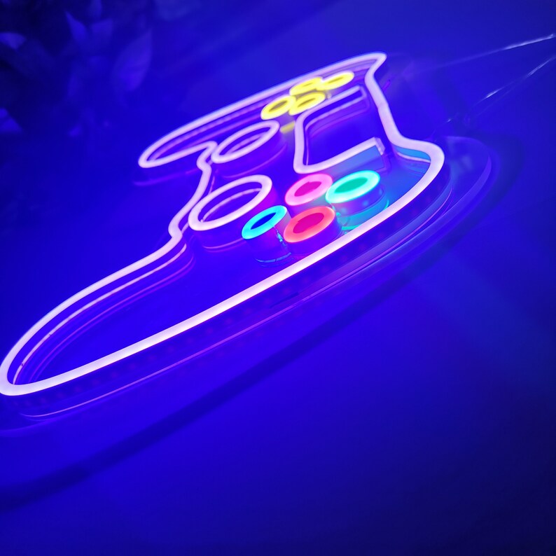 May include: A neon sign shaped like a video game controller with colorful buttons. The sign is lit up in a bright purple hue.