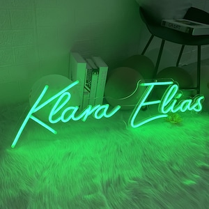 Puede incluir: Un letrero de ne&oacute;n verde con los nombres "Klara Elias" en una fuente cursiva. El letrero est&aacute; sobre una superficie verde y peluda.