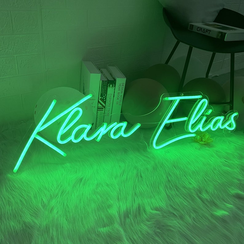Puede incluir: Un letrero de ne&oacute;n verde con los nombres "Klara Elias" en una fuente cursiva. El letrero est&aacute; sobre una superficie verde y peluda.