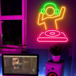 May include: Neon sign depicting a DJ with headphones, arms raised, and a turntable. The sign features a neon green outline for the headphones, a yellow outline for the DJ figure, and a pink outline for the turntable base. The sign is mounted on a wall.