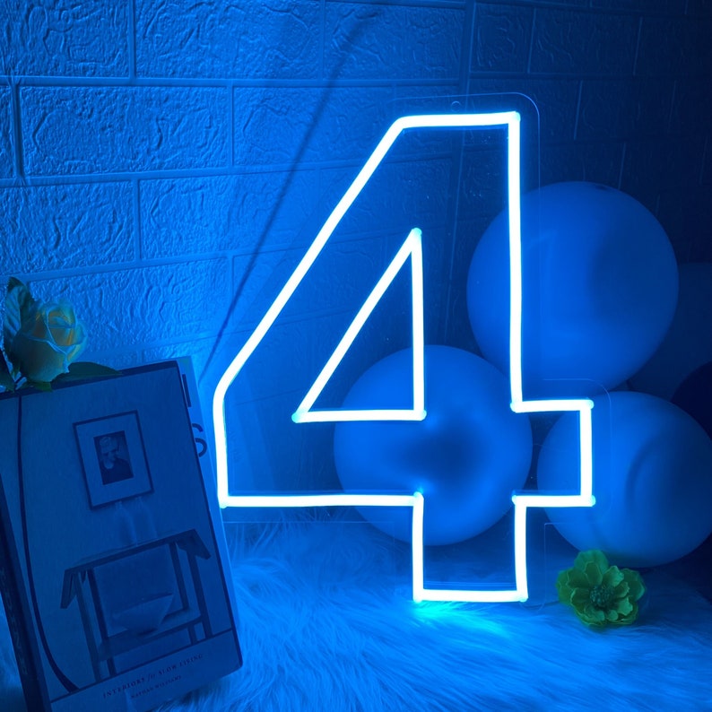 Pu&ograve; includere: Un cartello al neon bianco a forma di numero quattro, che brilla di blu. Il cartello &egrave; appoggiato su una superficie blu pelosa con palloncini bianchi dietro.