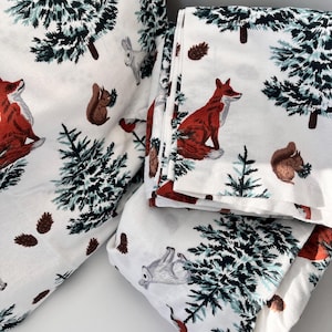Puede incluir: Ropa de cama blanca con un patrón de bosque que incluye zorros rojos, ardillas, conejos y pinos. La ropa de cama está hecha de tela suave y cómoda.