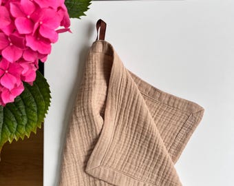 May include: A beige, textured towel with a stitched border hangs from a brown leather loop. The towel is folded, showing its corner. Bright pink hydrangea flowers and green leaves are in the background.
