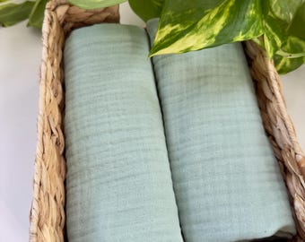 May include: Two rolled, light green muslin cloths are nestled in a woven wicker basket. The basket is rectangular with rounded corners. Green and yellow variegated leaves and vines are in the background.