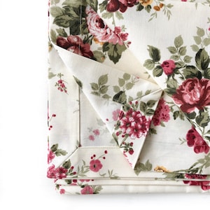 May include: Close-up of a folded fabric with a floral pattern. The fabric is cream-colored with pink and green roses and leaves.