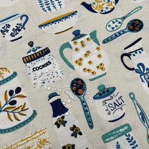 May include: A white fabric with a blue and yellow floral pattern. The fabric features a variety of kitchen items, including teapots, bowls, spoons, and jars. The fabric is water-resistant and has a subtle sheen.