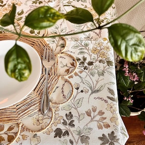 May include: A white tablecloth with a floral pattern in shades of green, brown, and yellow. A woven placemat with a scalloped edge and a silver fork are on top of the tablecloth.