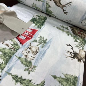 May include: White fabric with a winter scene print featuring deer, a red cabin, and pine trees. The fabric has a watercolour-like effect.