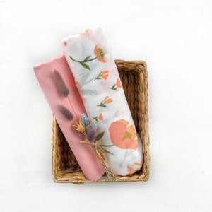 May include: Two rolled baby swaddles in a woven basket. One is solid pink, the other white with peach floral print. A small bundle of dried flowers is tied with twine. The basket is on a white surface.