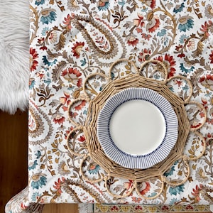 以下が含まれることがあります： 茶色、赤、青の色合いのペイズリー柄の白いテーブルクロス。テーブルクロスの上に、白いプレートに青いストライプが入った籐製のプレースマットが置かれています。