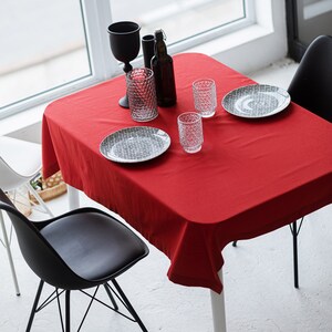 Puede incluir: Un mantel rojo cubre una mesa blanca con sillas negras. La mesa está puesta con dos platos, dos vasos, una botella y dos vasos.