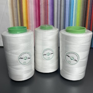 Può includere: Tre grandi rocche di filo da cucito bianco 100% poliestere. Ogni rocca presenta una parte superiore in plastica verde brillante e un'etichetta circolare bianca con il testo Bella Textile, 100% polyester sewing thread, e 40/2 stampato in nero.