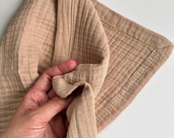 May include: A beige muslin blanket featuring a soft, crinkled texture and a stitched border. The fabric has a lightweight, multi-layered appearance with a grid-like pattern woven into the material, showcasing a neutral, earthy tone throughout.