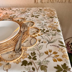 May include: A white tablecloth with a floral pattern in shades of green, yellow, and brown. A woven placemat with a white plate and a silver fork are on the tablecloth.