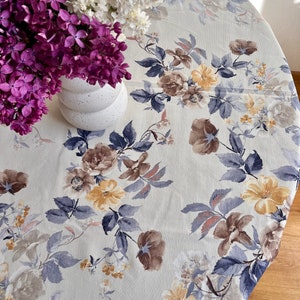 Könnte beinhalten: Eine weiße Tischdecke mit einem floralen Muster in Blautönen, Braun und Gelb. Die Tischdecke ist rund und hat eine strukturierte Oberfläche. Ein Strauß aus lila und weißen Blumen steht in einer weißen Vase auf der Tischdecke.